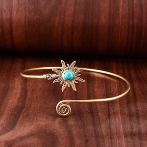 May include: A gold-colored arm cuff featuring a sunburst design with a turquoise center stone. The cuff has a spiral detail at one end and is displayed on a wooden surface. This piece of jewelry is a unique accessory.