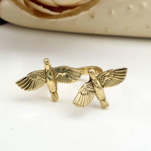 Puede incluir: Un anillo dorado con dos esculturas detalladas de pájaros con las alas extendidas. Los pájaros están colocados a ambos lados de la banda del anillo, creando un diseño simétrico. El anillo está sobre un fondo blanco.