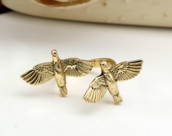 May include: A gold-coloured ring featuring two detailed bird sculptures with outstretched wings. The birds are positioned on opposite sides of the ring band, creating a symmetrical design. The ring is set against a white background.