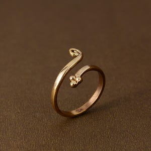 May include: A gold-colored ring with an open design. The ring features a curved band that wraps around the finger, with a small cluster of three beads and a swirl detail. The ring is set against a brown background.