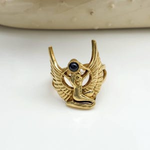 Op de afbeelding: Goudkleurige ring met een gevleugeld ontwerp, een centrale figuur en een donkere, cirkelvormige steen. De vleugels van de ring hebben veermotieven en de figuur lijkt te knielen. De ring staat tegen een witte achtergrond.
