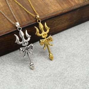Puede incluir: Dos collares con colgantes de tridente. Un colgante es plateado, el otro es dorado. Ambos colgantes tienen un diseño en forma de lazo debajo del tridente. Los collares están sobre una superficie de madera.