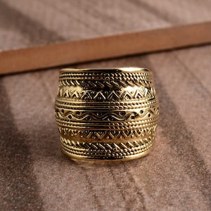 Könnte beinhalten: Ein goldfarbener Ring mit einem breiten Band, das mit einem komplizierten geometrischen Muster verziert ist. Der Ring hat eine strukturierte Oberfläche und eine leicht abgerundete Form.