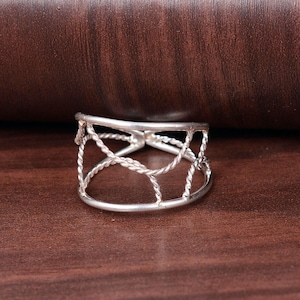 May include: A silver ring with a unique open design. The ring features a series of interconnected, twisted rope-like patterns that form a cage-like structure around the finger. The ring is set against a dark brown wooden surface.