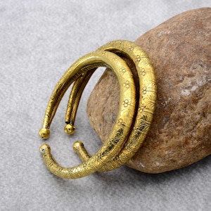 Op de afbeelding: Twee goudkleurige, open armbanden met ingewikkelde bloemen- en bladerpatronen. Elke armband heeft een afgerond, bolvormig uiteinde. De armbanden liggen op een stenen oppervlak.