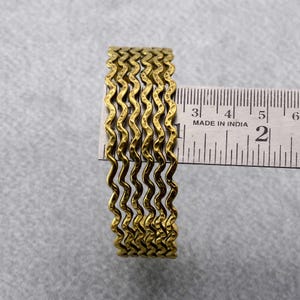 Può includere: Sei braccialetti in metallo dorato con un design ondulato e testurizzato. I braccialetti sono impilati e misurati con un righello, mostrando che hanno un diametro di circa 5 cm.