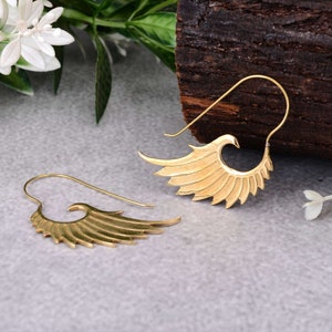 Może przedstawiać: Para złotych kolczyków w kształcie skrzydeł. Kolczyki mają haczykowaty kształt do przekłutych uszu. Skrzydła są szczegółowe z wieloma liniami przypominającymi pióra. Kolczyki są prezentowane na szarej powierzchni z drewnianym tłem.