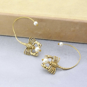 May include: A pair of gold-tone scorpion earrings with pearl accents. The earrings are made of a thin wire that wraps around the ear and has a scorpion charm attached to it.