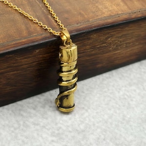 Puede incluir: Collar con colgante dorado con un diseño cilíndrico con una serpiente enrollada. El colgante cuelga de una delicada cadena dorada. El collar está sobre un fondo de madera.
