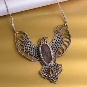 May include: A handmade macrame necklace featuring a central oval stone in shades of brown and green. The gray macrame design is shaped like a bird with outstretched wings, accented with small gold beads. The necklace hangs on a gray cord.