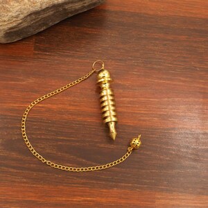 Puede incluir: Un péndulo dorado con una cadena y una bola decorativa en el extremo. El péndulo tiene un cuerpo cilíndrico con estrías horizontales y una punta. La cadena está unida a la parte superior del péndulo.