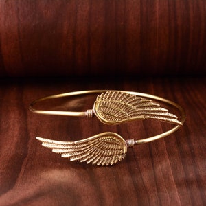 May include: A gold-toned bangle bracelet with two wing-shaped charms. The wings are intricately detailed and have a delicate, feminine feel.