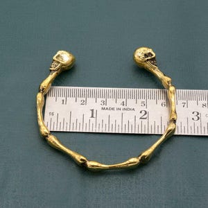 May include: Gold-toned bracelet with two skull-shaped ends. The bracelet is measured against a ruler that reads "Made in India".