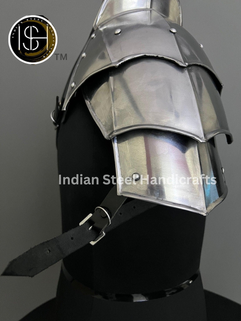 May include: A silver metal shoulder armour with a layered design and a black leather strap. The armour is displayed on a black mannequin.