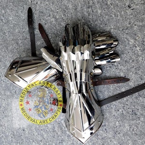 May include: A pair of silver plated medieval gauntlets with leather straps. The gauntlets are made of metal plates and have a pointed design. The gauntlets are on a gray surface.  MEDIEVAL ART CRAFTS is printed on a circular label.