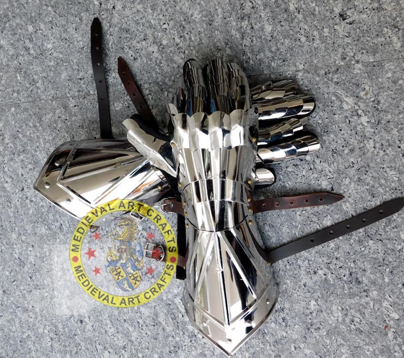 May include: A pair of silver plated medieval gauntlets with leather straps. The gauntlets are made of metal plates and have a pointed design. The gauntlets are on a gray surface.  MEDIEVAL ART CRAFTS is printed on a circular label.