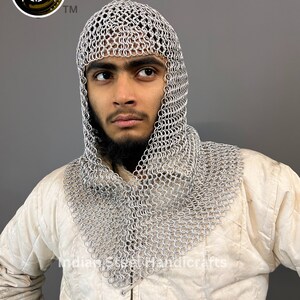 May include: A silver chainmail coif with a hood, worn by a person with a beard. The coif is made of interlocking metal rings and covers the head and neck.