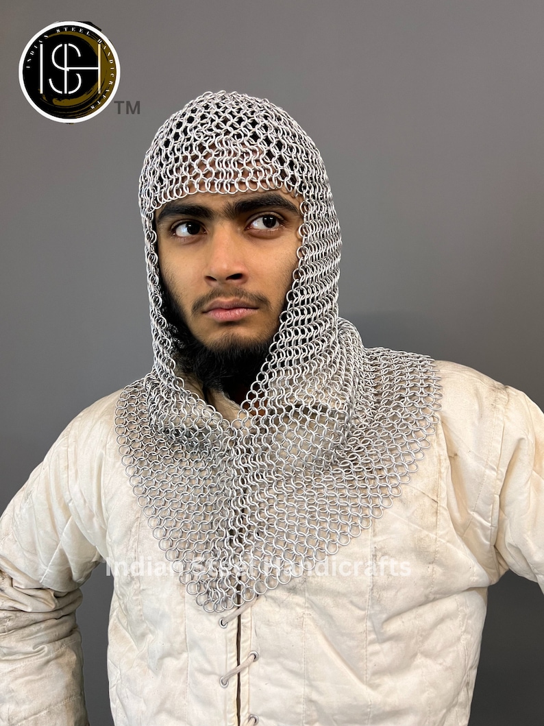 May include: A silver chainmail coif with a hood, worn by a person with a beard. The coif is made of interlocking metal rings and covers the head and neck.