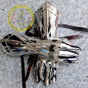 May include: A pair of silver plated medieval gauntlets with a riveted design. The gauntlets are made of metal and have a polished finish. The gauntlets are attached to leather straps.