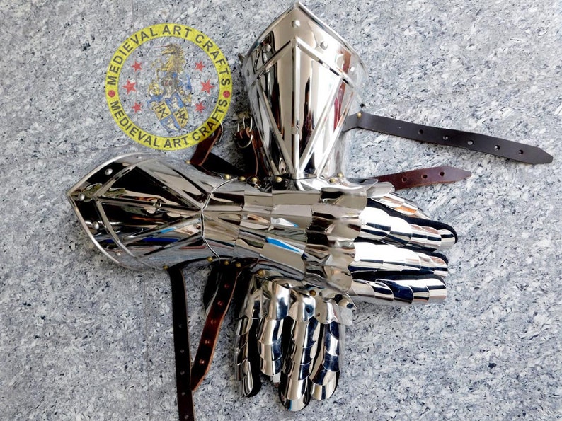 May include: A pair of silver plated medieval gauntlets with a riveted design. The gauntlets are made of metal and have a polished finish. The gauntlets are attached to leather straps.