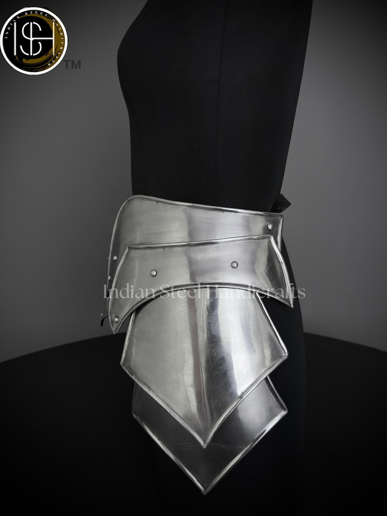 May include: Silver metal armor for the lower torso, with three overlapping plates, worn on a black mannequin. The armor is shaped like a series of overlapping triangles, with a curved top plate. The text "Indian Steel Handcrafts" is visible on the armor.