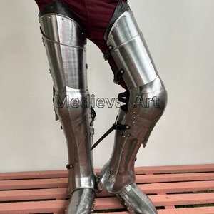 May include: A pair of silver-colored metal leg armor with pointed toes. The armor is worn over brown pants and a black belt. The armor is made of metal plates and has a decorative design.