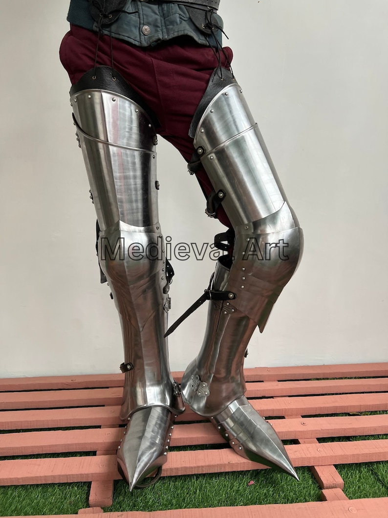 May include: A pair of silver-colored metal leg armor with pointed toes. The armor is worn over brown pants and a black belt. The armor is made of metal plates and has a decorative design.