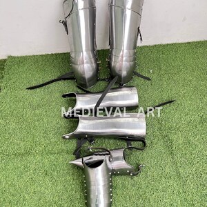 May include: A set of medieval leg armor, including shin guards, knee guards, and greaves. The armor is made of polished steel and has a silver finish. The armor is laid out on a green grassy surface.