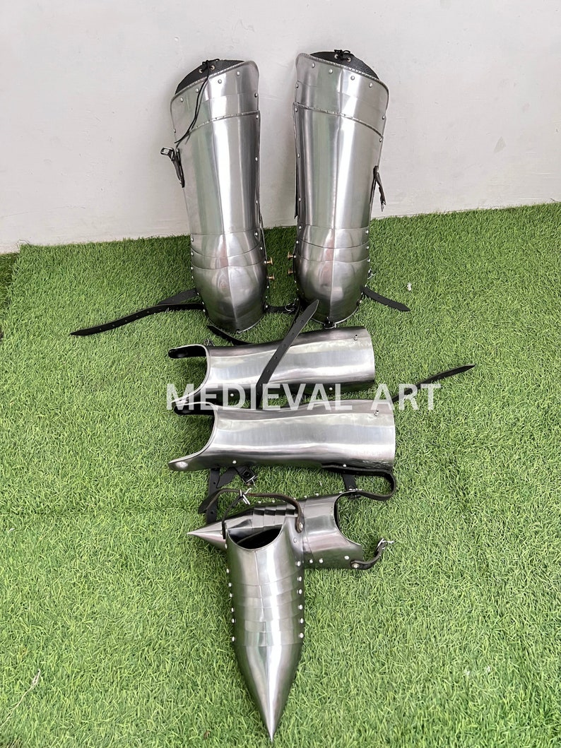 May include: A set of medieval leg armor, including shin guards, knee guards, and greaves. The armor is made of polished steel and has a silver finish. The armor is laid out on a green grassy surface.