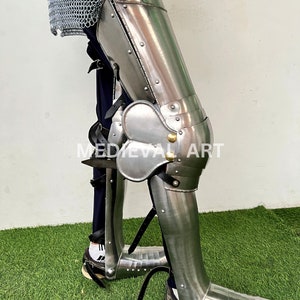 May include: A silver metal leg armor with a knee pad and a pointed toe. The armor is worn over a pair of black pants and a chainmail shirt. The armor is secured with straps and buckles. The armor is on a person's leg and is standing on a green grassy surface.  MEDIEVAL ART is printed on the armor.