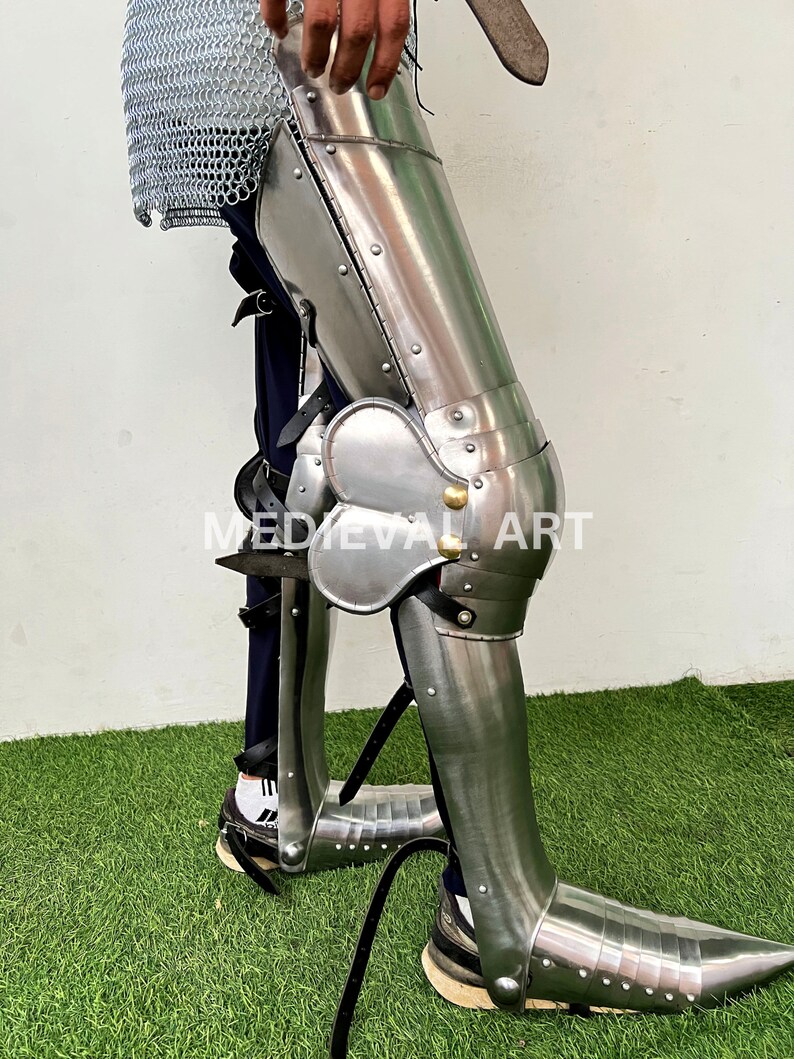 May include: A silver metal leg armor with a knee pad and a pointed toe. The armor is worn over a pair of black pants and a chainmail shirt. The armor is secured with straps and buckles. The armor is on a person's leg and is standing on a green grassy surface.  MEDIEVAL ART is printed on the armor.