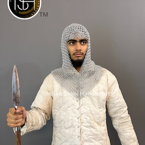 May include: A person wearing a white quilted jacket and a silver chainmail coif, holding a wooden-handled spear with a metal tip. The person is looking to the right. The background is a grey wall. The image also includes a logo with the text "Indian Steel Handicrafts TM".