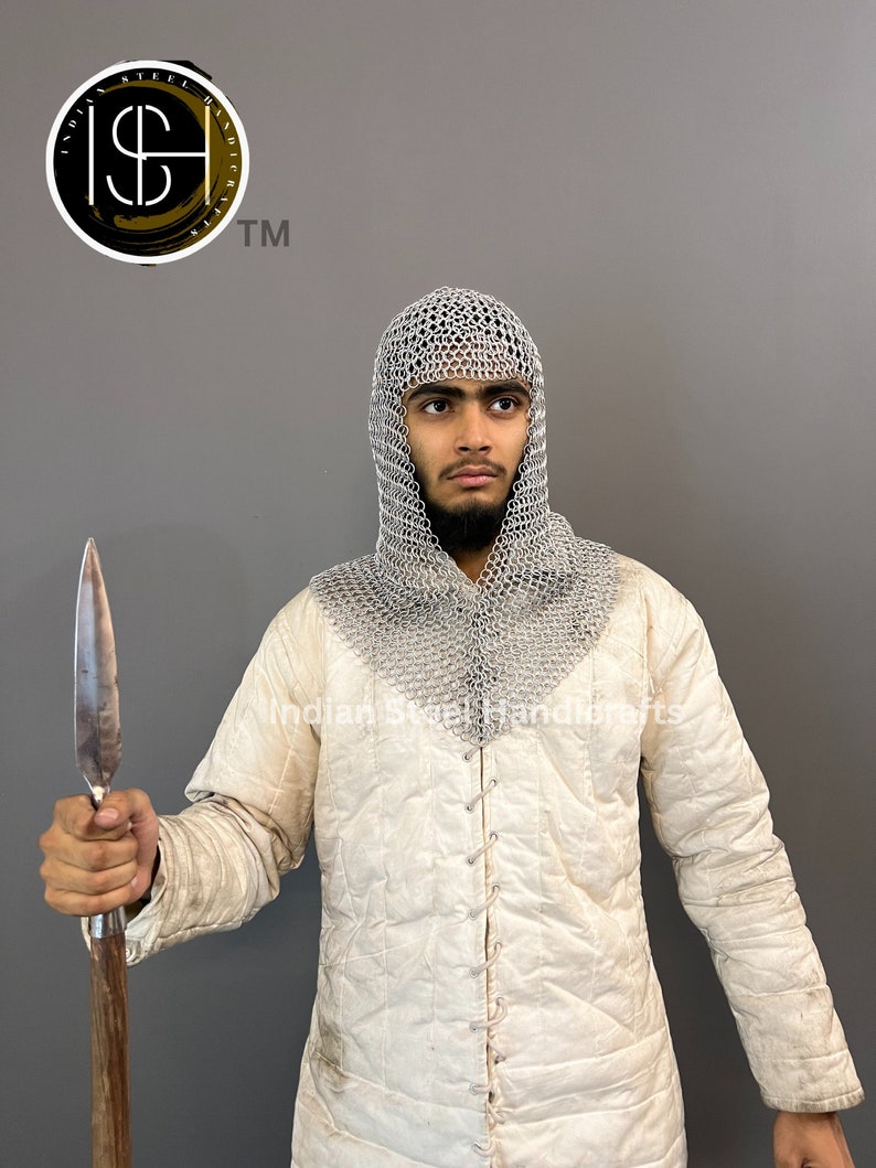 May include: A person wearing a white quilted jacket and a silver chainmail coif, holding a wooden-handled spear with a metal tip. The person is looking to the right. The background is a grey wall. The image also includes a logo with the text "Indian Steel Handicrafts TM".