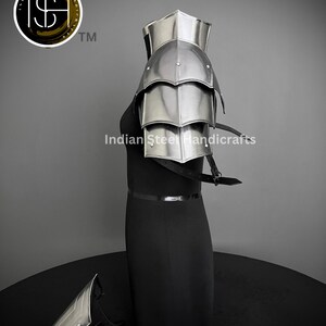 K&ouml;nnte beinhalten: Eine silberne Schulterr&uuml;stung aus Metall mit einem geschichteten Design. Die R&uuml;stung befindet sich auf einem schwarzen Mannequin. Die R&uuml;stung besteht aus mehreren Platten, die durch Nieten verbunden sind. Die R&uuml;stung wird am rechten Arm getragen. Die R&uuml;stung ist Teil eines Sets, das eine passende Unterarmr&uuml;stung umfasst. Die Unterarmr&uuml;stung liegt auf dem Boden vor dem Mannequin. Das Bild enth&auml;lt auch den Text "Indian Steel Handicrafts" und ein Logo mit den Buchstaben "ISH" und einem Dollarzeichen.
