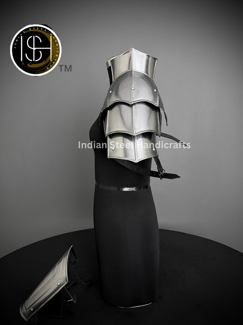 K&ouml;nnte beinhalten: Eine silberne Schulterr&uuml;stung aus Metall mit einem geschichteten Design. Die R&uuml;stung befindet sich auf einem schwarzen Mannequin. Die R&uuml;stung besteht aus mehreren Platten, die durch Nieten verbunden sind. Die R&uuml;stung wird am rechten Arm getragen. Die R&uuml;stung ist Teil eines Sets, das eine passende Unterarmr&uuml;stung umfasst. Die Unterarmr&uuml;stung liegt auf dem Boden vor dem Mannequin. Das Bild enth&auml;lt auch den Text "Indian Steel Handicrafts" und ein Logo mit den Buchstaben "ISH" und einem Dollarzeichen.
