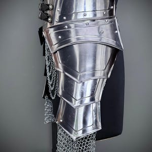 K&ouml;nnte beinhalten: Poliertes Metall-R&uuml;stungsteil, wahrscheinlich f&uuml;r Bein und H&uuml;fte, mit geschichteten Platten und Kettengeflecht-Akzenten. Die R&uuml;stung wird auf einer schwarzen Form vor grauem Hintergrund pr&auml;sentiert. Nieten und Riemen sichern die Platten.