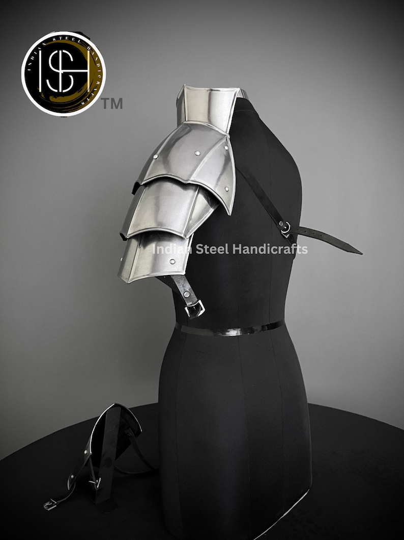 K&ouml;nnte beinhalten: Silberbeschichteter Schulterpanzer aus Metall mit einem mehrschichtigen Design. Die R&uuml;stung ist an einem schwarzen Mannequin mit einem schwarzen Lederriemen befestigt. Der Text "Indian Steel Handicrafts" ist auf dem Mannequin sichtbar.