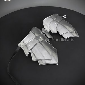 May include: A pair of silver metal arm guards with a black leather strap. The guards are made of multiple overlapping plates and have a riveted design. The text "Indian Steel Handicrafts" is visible on the guards.