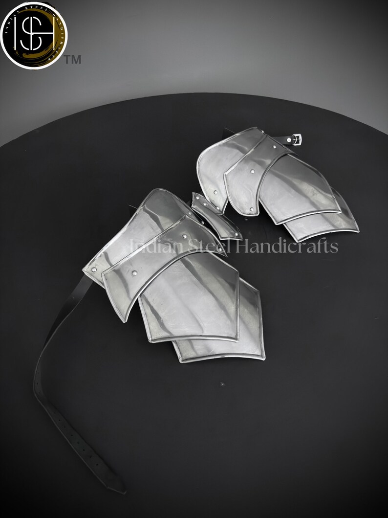 May include: A pair of silver metal arm guards with a black leather strap. The guards are made of multiple overlapping plates and have a riveted design. The text "Indian Steel Handicrafts" is visible on the guards.