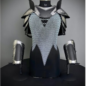 May include: A suit of armor displayed on a black mannequin. The armor includes a metal chest piece with chainmail, shoulder guards, and arm guards. The armor is silver and black, and is displayed against a gray background.