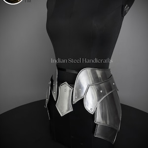 May include: Silver metal armor skirt with a decorative design. The skirt is made of multiple plates that are connected by rivets. The skirt is worn on a black mannequin. The text "Indian Steel Handicrafts" is visible on the mannequin.