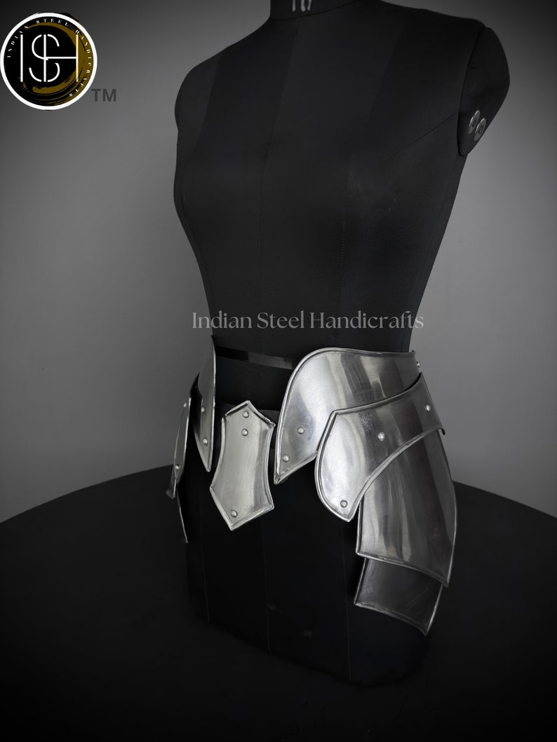 May include: Silver metal armor skirt with a decorative design. The skirt is made of multiple plates that are connected by rivets. The skirt is worn on a black mannequin. The text "Indian Steel Handicrafts" is visible on the mannequin.