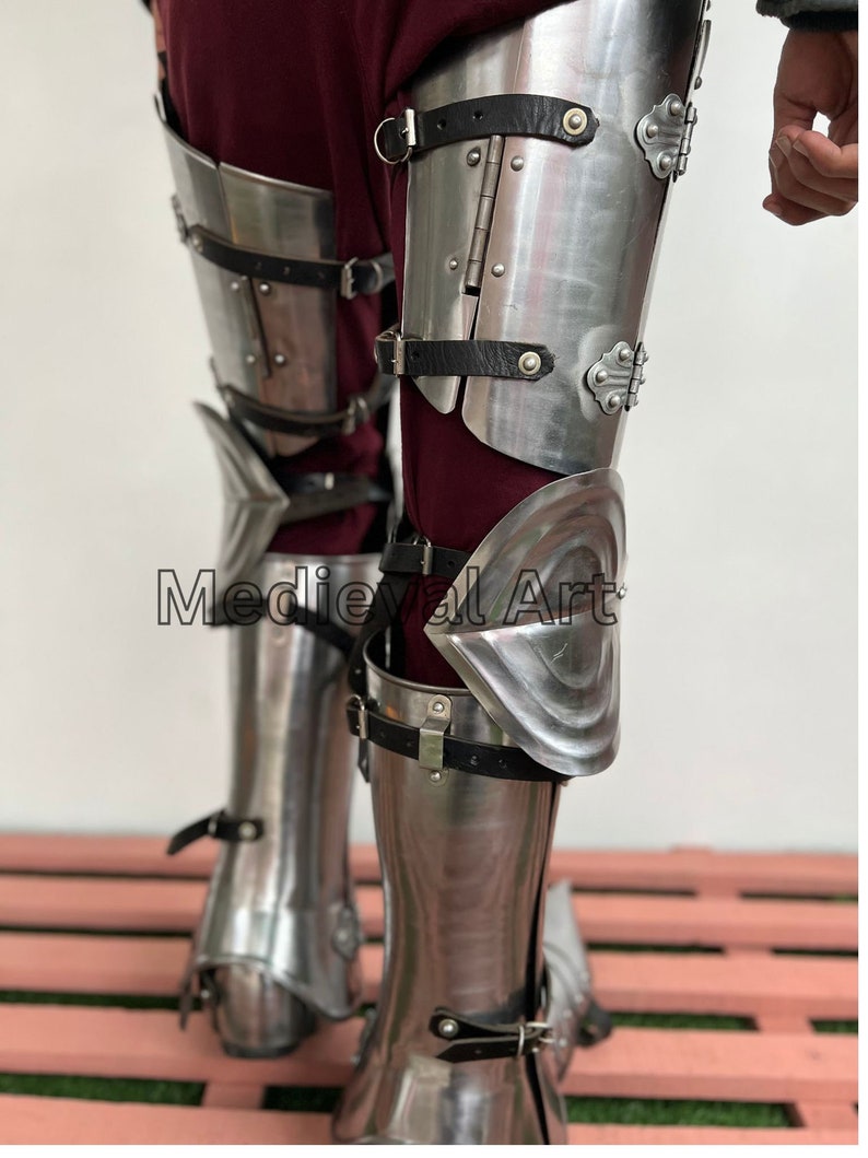 May include: A pair of silver metal leg armor with black leather straps and buckles. The armor is designed to protect the shins, knees, and thighs. The armor is worn over a pair of brown pants.