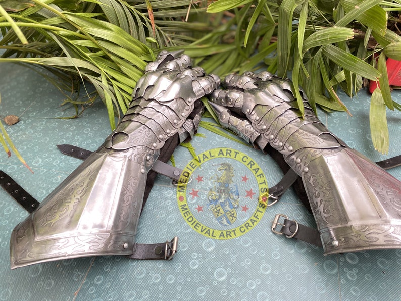 Puede incluir: Un par de guanteletes medievales plateados con detalles intrincados. Los guanteletes est&aacute;n hechos de placas de metal y presentan dedos articulados. Correas de cuero negro con hebillas aseguran los guanteletes. El fondo muestra follaje verde.