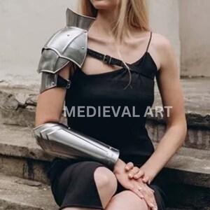 K&ouml;nnte beinhalten: Ein silberner Armschutz aus Metall mit passendem Unterarmschutz. Der Armschutz hat ein dekoratives Design und wird am linken Arm getragen. Der Unterarmschutz wird am linken Unterarm getragen. Der Armschutz ist mit einem Riemen am Unterarmschutz befestigt.