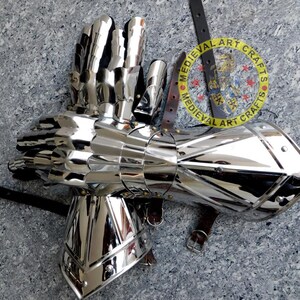 May include: A pair of silver plated medieval gauntlets with a segmented design. The gauntlets are made of metal and have a polished finish. The gauntlets are attached to a leather strap with a buckle. The strap has a circular logo with the text "MEDIEVAL ART CRAFTS".