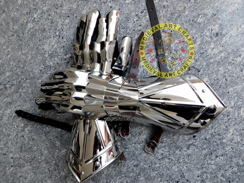 May include: A pair of silver plated medieval gauntlets with a segmented design. The gauntlets are made of metal and have a polished finish. The gauntlets are attached to a leather strap with a buckle. The strap has a circular logo with the text "MEDIEVAL ART CRAFTS".