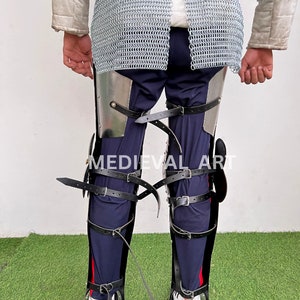 May include: Black leather leg armor with metal plates and buckles. The armor is worn over blue pants and secured with straps. The text "MEDIEVAL ART" is visible on the armor.