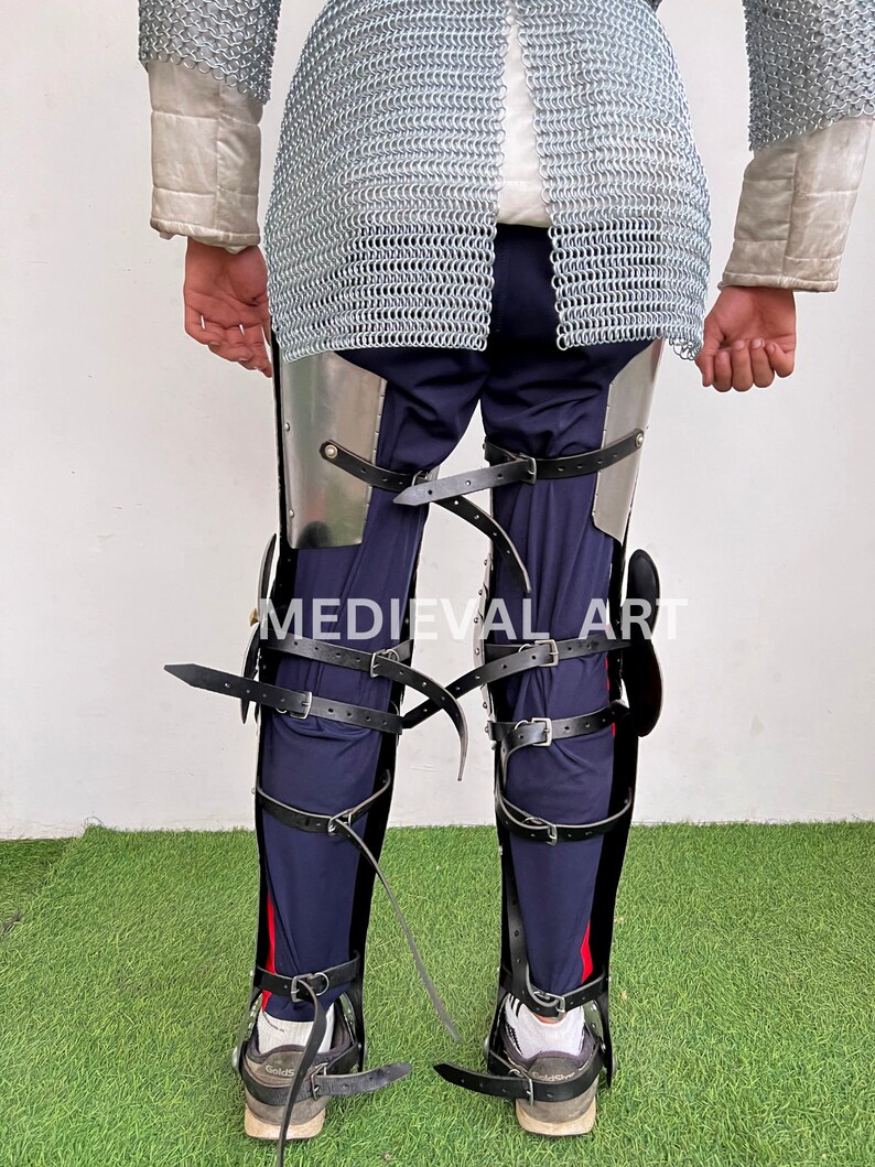 May include: Black leather leg armor with metal plates and buckles. The armor is worn over blue pants and secured with straps. The text "MEDIEVAL ART" is visible on the armor.
