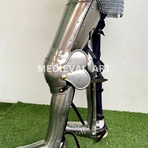 May include: A pair of silver metal leg armor with a pointed toe, knee pads, and straps. The armor is worn over blue pants and white socks.